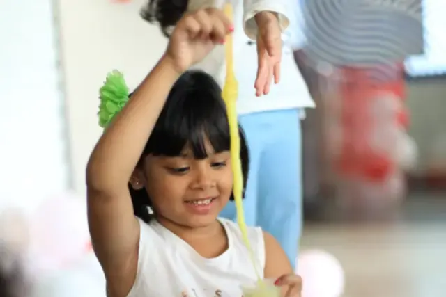 A young girl grinning with one arm raised, holding a small tub of slime at a birthday party