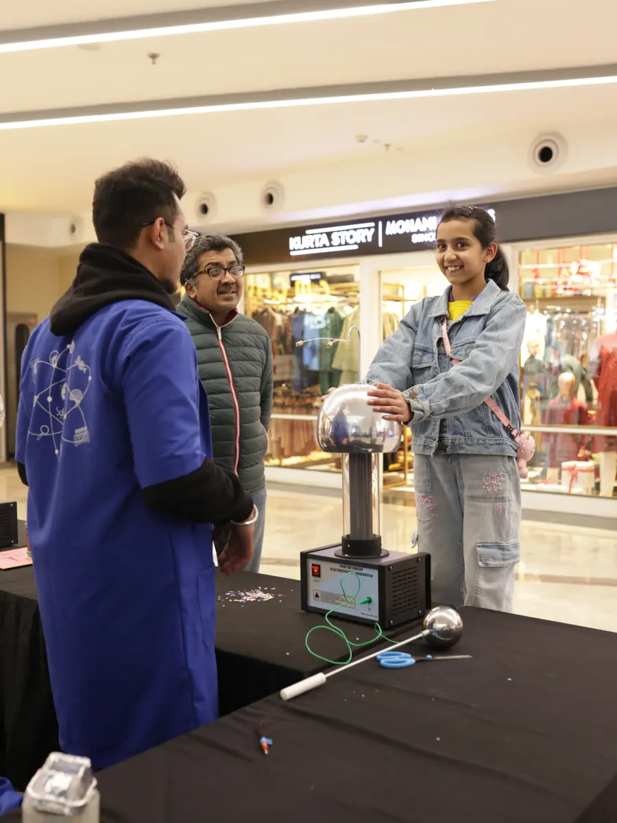 A girl resting both hands on a Van de Graaff generator while a MakerMasti host explains what is happening