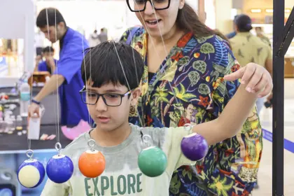 A MakerMasti host swinging a row of coloured pendulum balls while a boy in glasses tracks the motion